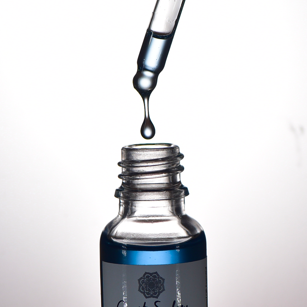 Close-up capturing the moment an oil droplet falls into the 'Clear Skies' face serum bottle as a dropper hovers above the open bottle, releasing a luxurious blue oil.