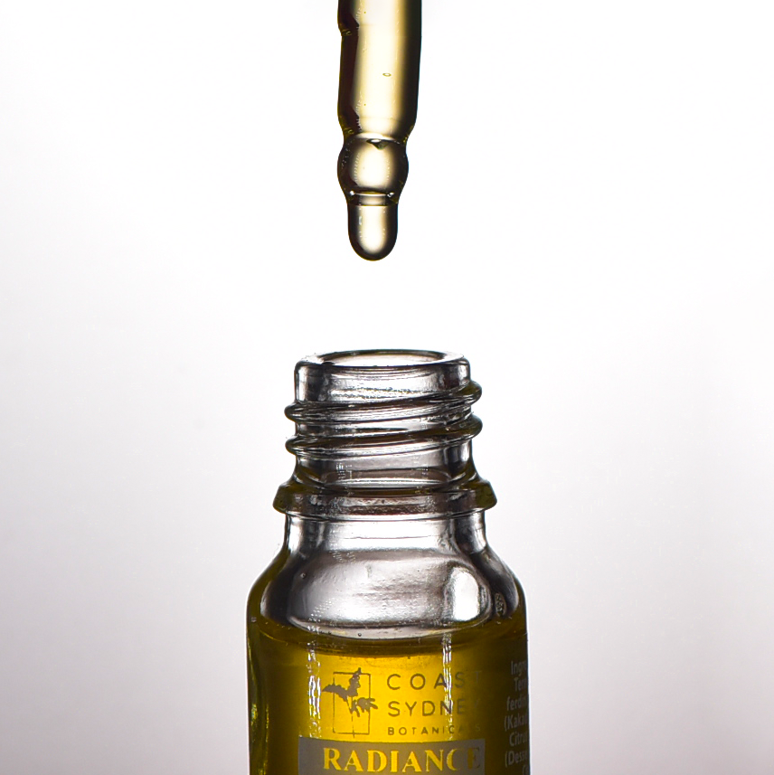 Close-up capturing the moment an oil droplet falls into the 'Radiance' face serum bottle as a dropper hovers above the open bottle, releasing a luxurious yellow oil.
