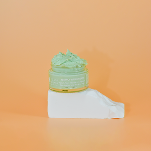 "simply stronger' green moisturiser on an orange background, displayed on a white stone block. The jar's open lid reveals the creamy texture of the moisturiser